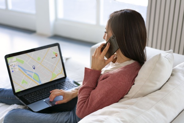 Photo d'une femme assise dans son canapé, dans son salon. Elle est en train d'appeler quelqu'un et regarde, posé sur ses genoux, son ordinateur portable sur lequel on voit un résultat de recherche sur Google Maps. Elle semble avoir trouvé un prestataire local, près de chez elle. 